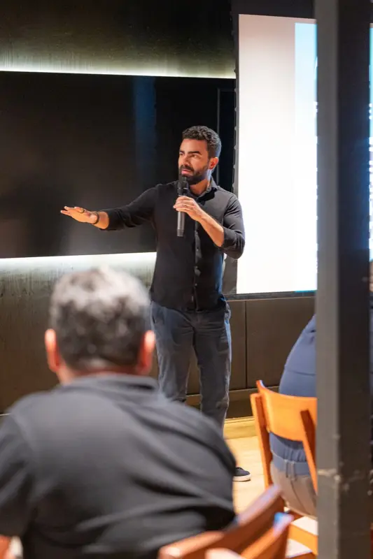 Palestrante no palco se dirige ao público, que aparece de costas em primeiro plano.
