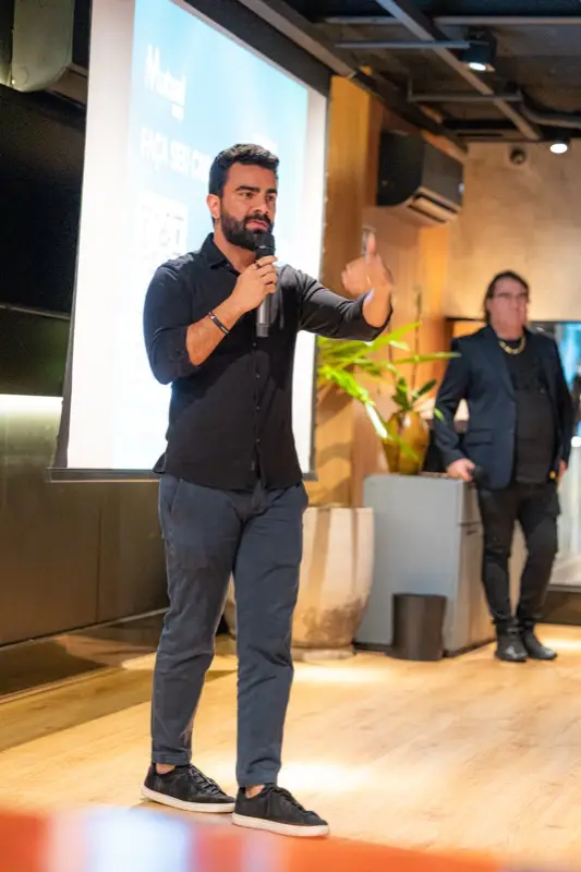 Palestrante no palco fala ao microfone, com o telão ao fundo.