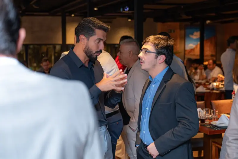 Dois participantes conversam no salão, com as mesas do encontro ao fundo.