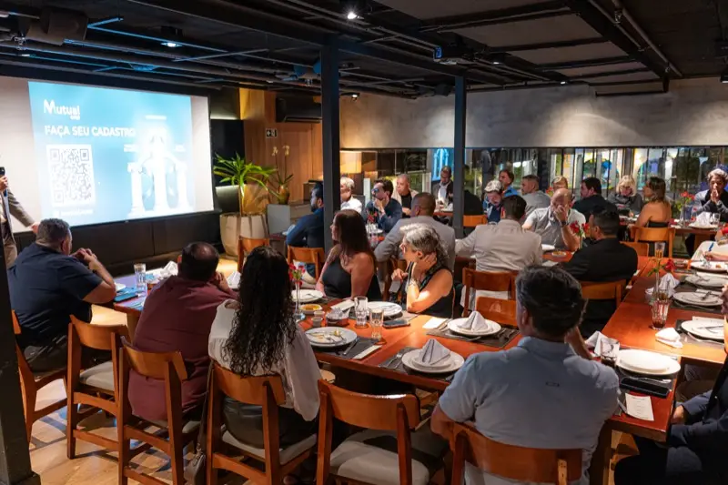 Salão cheio, com participantes sentados em mesas longas diante do telão que exibe o convite para fazer o cadastro na Mutualcoop.